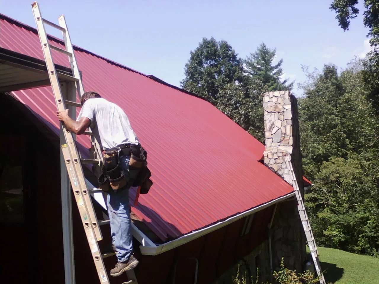 Roofer on a ladder beside a red metal roof and stone chimney — Soffit & Fascia Repair by Roofing Brothers in East Greenwich